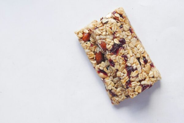 Two chewy granola bars made with rolled oats, almonds, cashews and dried cranberries on a pale grey surface