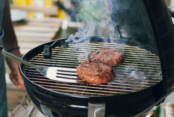 Two burger patties cooking on a charcoal grill as a cook lifts one with a metal spatula, smoke rising from the grate