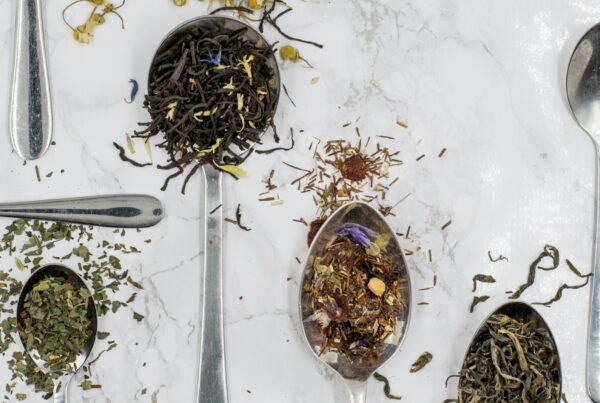 Loose leaf black, green, and mint teas and a dried fruit blend on silver spoons, scattered with botanicals and flower petals