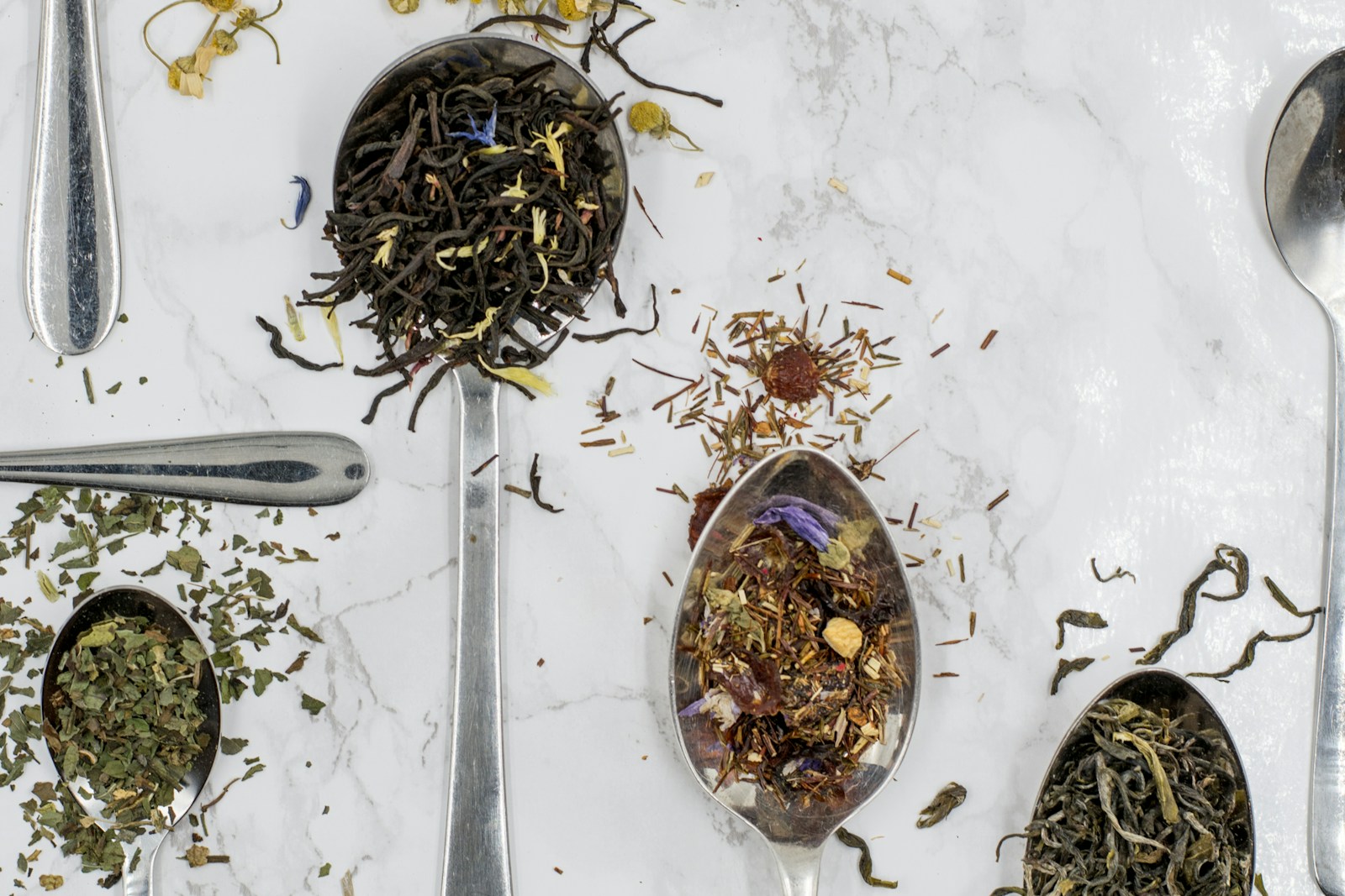 Loose leaf black, green, and mint teas and a dried fruit blend on silver spoons, scattered with botanicals and flower petals