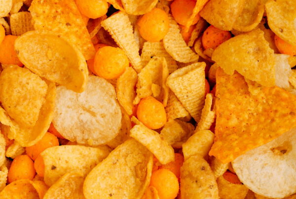 A close-up mix of seasoned salty snacks - tortilla chips, potato crisps, extruded corn cones and cheese puffs - showing the range of flavour coatings applied to snack products.