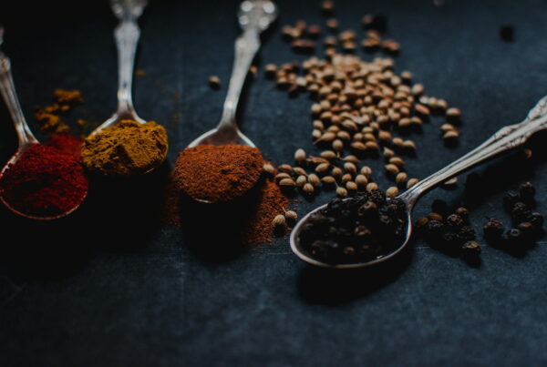 Spoons of ground chili, turmeric, and cinnamon beside coriander seed and black peppercorns on a dark surface