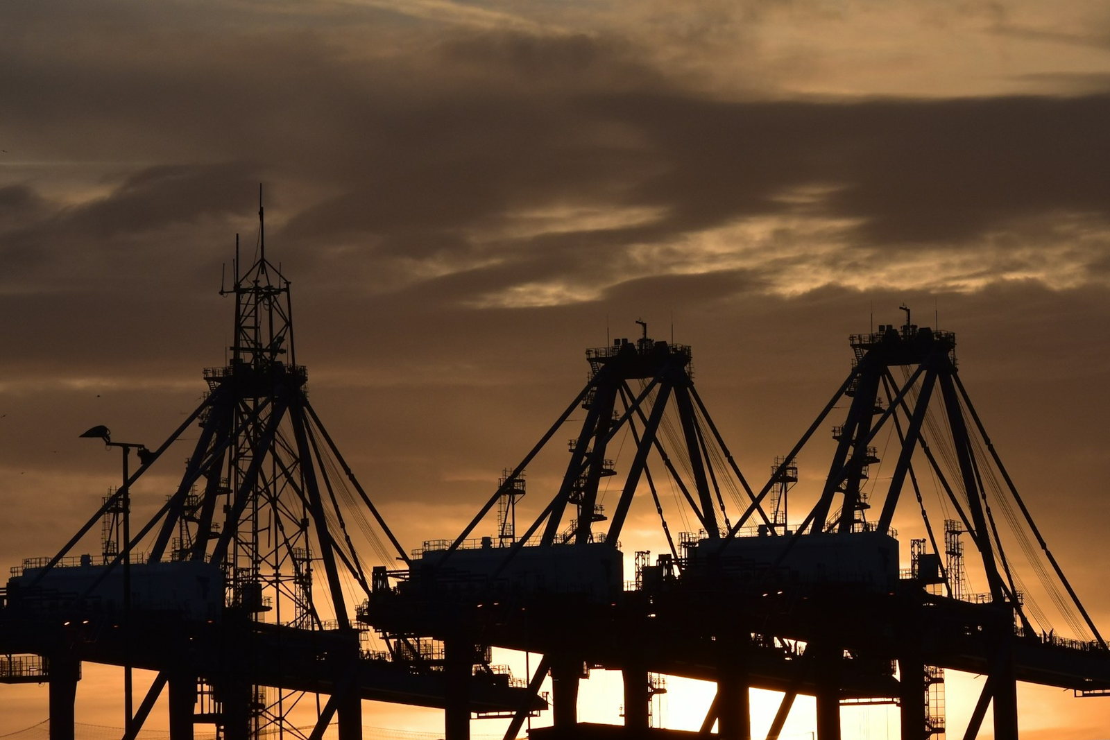 Three ship-to-shore container gantry cranes at a cargo port, silhouetted against an amber sunset sky