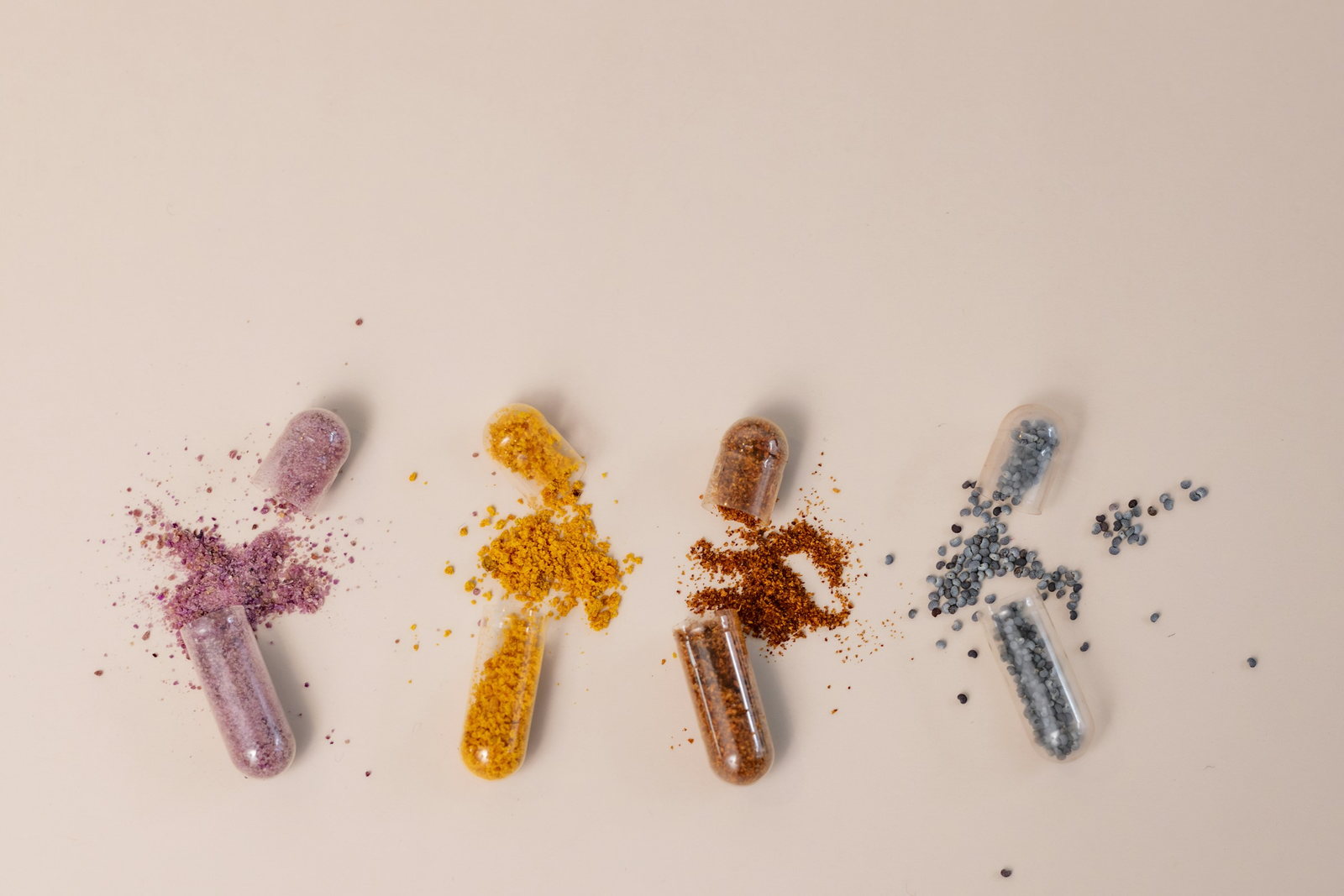 Four clear supplement capsules opened on a beige surface, each spilling a different coloured powder: purple, turmeric yellow, rust brown, and grey